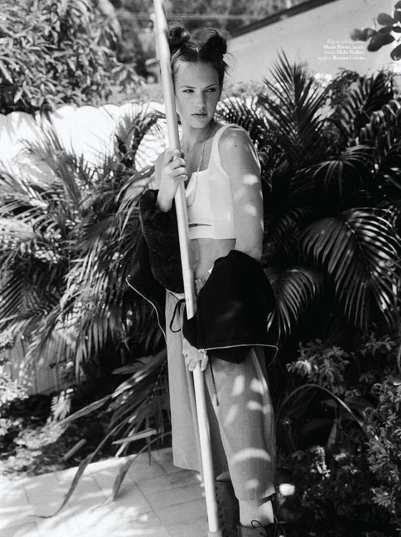 Black and white shot of a woman holding a pole, standing in a tropical garden with palm trees, wearing a crop top and baggy pants.