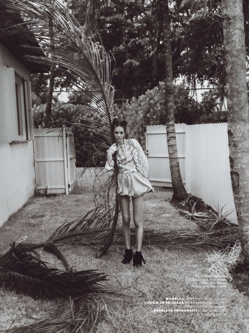 Monochrome fashion shot of a young woman posing in a ruffled top and shorts outdoors with palm fronds and a house in the background.