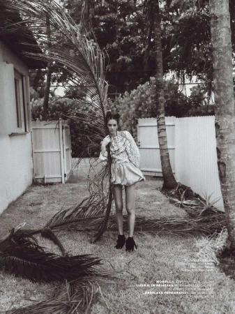 Monochrome fashion shot of a young woman posing in a ruffled top and shorts outdoors with palm fronds and a house in the background.