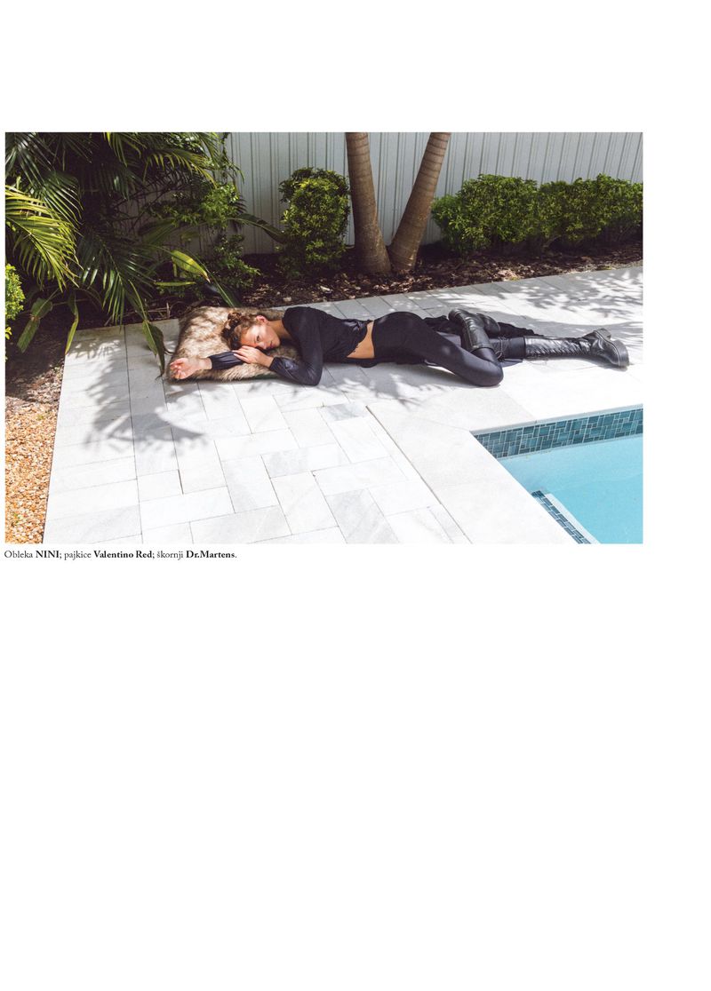 A young adult woman lies on a pillow on a tiled patio next to a pool in a black top and pants outdoors during daytime.