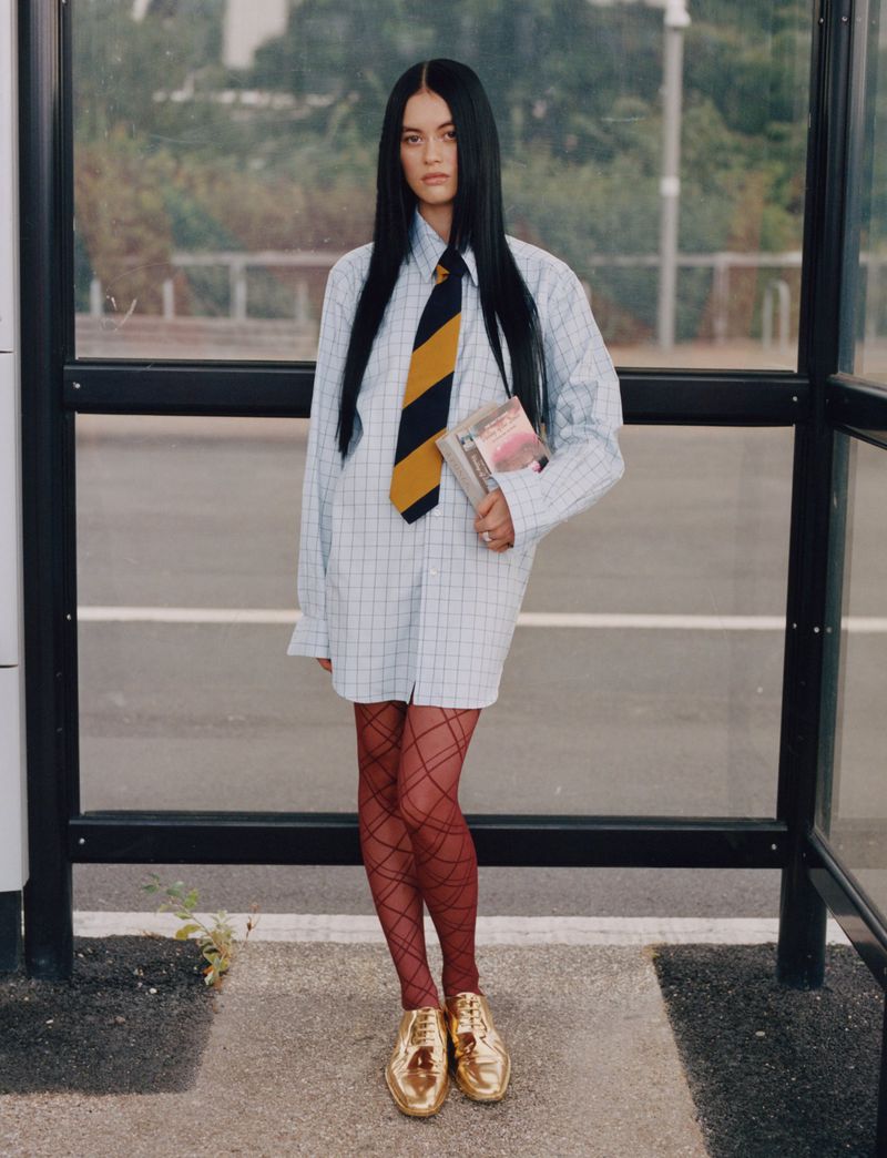 Young woman in a bus shelter, wearing an oversized shirt, tie, patterned tights and metallic gold shoes holds a book and looks at the camera.