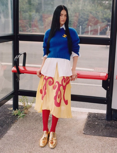 A young woman with long dark hair poses at a bus stop, wearing a blue sweater, white button-down shirt, yellow skirt, red tights, and gold shoes.