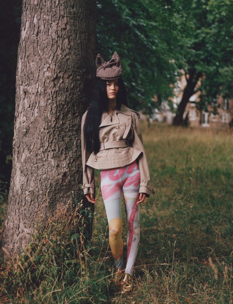 A young woman with long dark hair stands next to a tree in a grassy field wearing a tan jacket, patterned leggings, and a unique knitted hat.