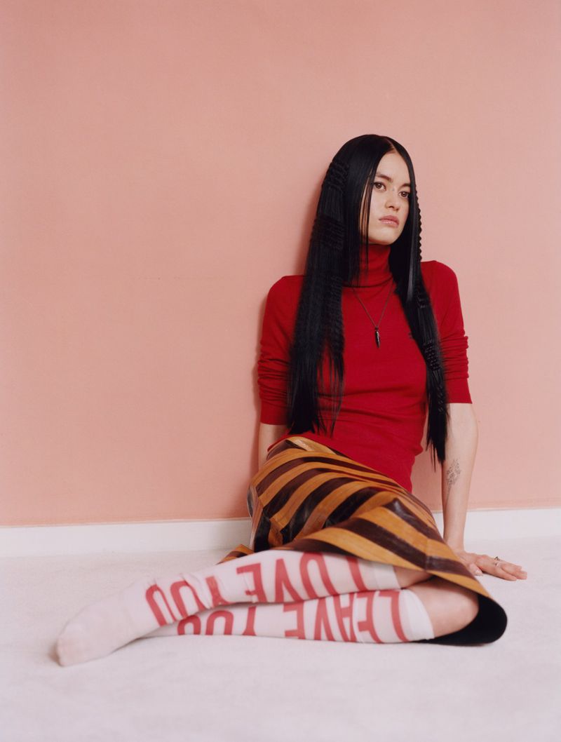 Portrait of a woman in a red turtleneck and brown-striped skirt sitting against a pink wall with knee-high socks with 'love you'.