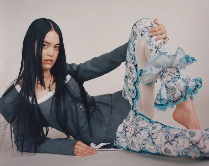 A fair-skinned woman with long dark hair poses in a studio, wearing a grey top and lacy blue and white pants, lying on the ground.