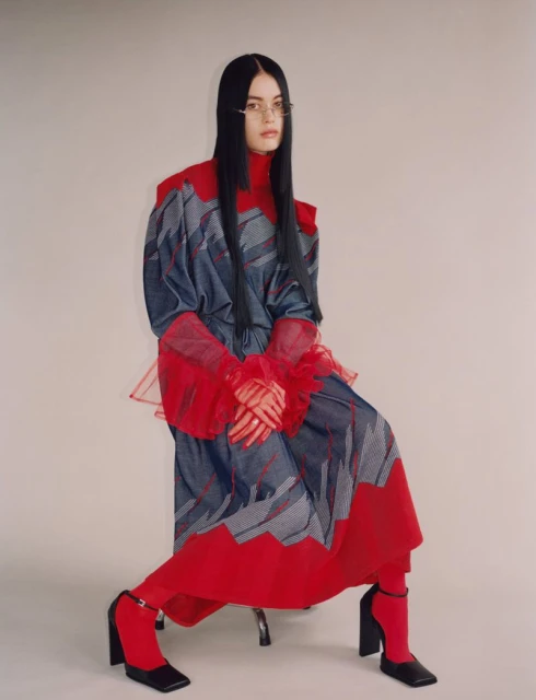 A woman with long black hair poses in an avant-garde, oversized dress with a red collar and cuffs in a studio setting. She wears black heels.