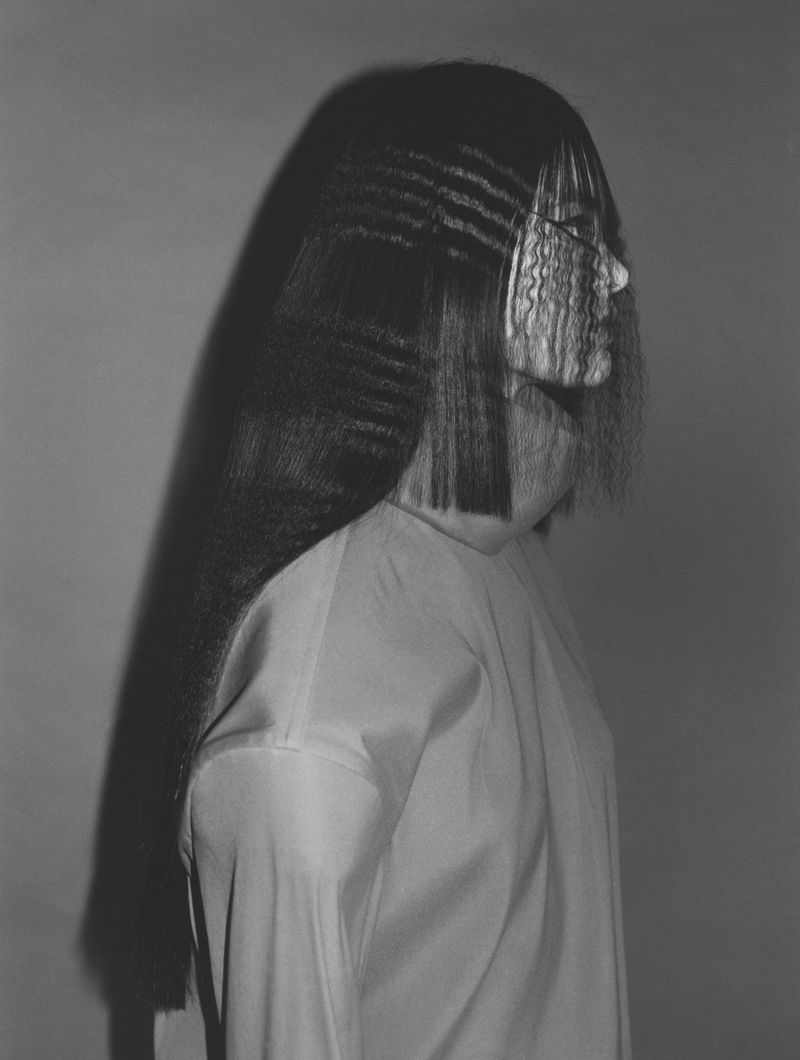 Monochrome profile of an adult woman with long, crimped black hair and a white shirt against a gray background in a studio setting.