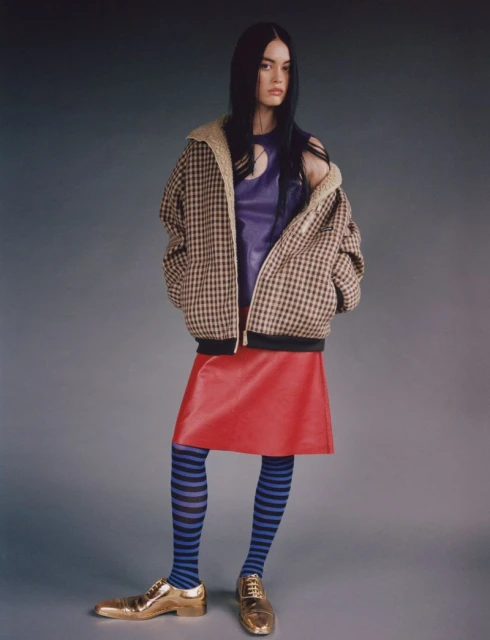 A full-length shot of a woman in a purple top, red skirt, and patterned tights poses against a gray background in a studio setting.