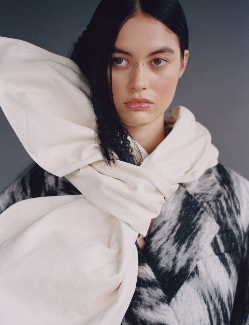Model Mimi Dey wearing a voluminous cream neck wrap and a textured black and grey coat against a neutral grey studio backdrop.
