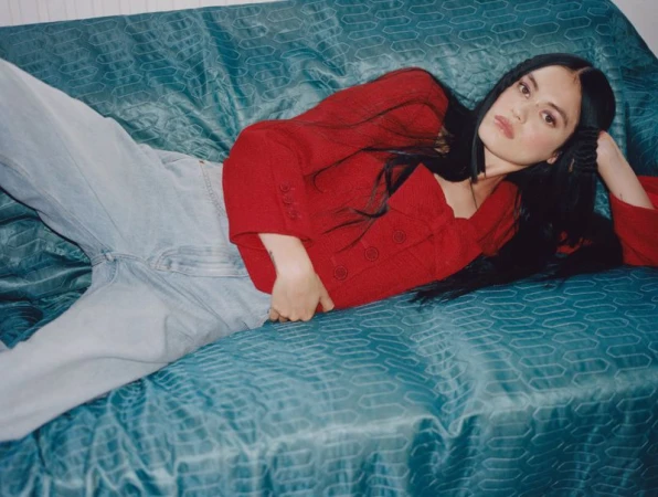 Brunette woman with a red jacket and jeans lying on a patterned blue sofa. The indoor studio shot is a medium shot, eye-level angle.