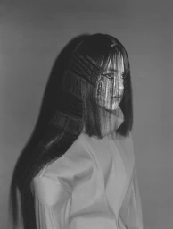 Black and white studio portrait of model Mimi Dey with sculptural layered hair wearing a high-collar coat in profile view.
