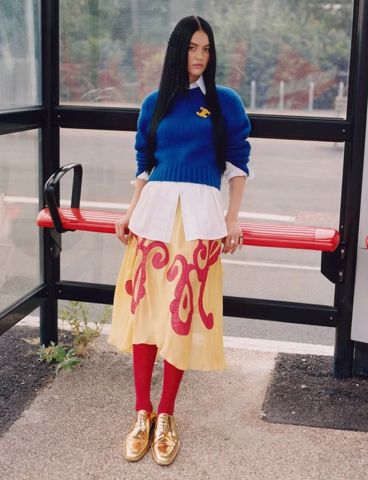Model with long dark hair wearing a blue sweater, white shirt, yellow patterned skirt, red tights, and gold shoes.