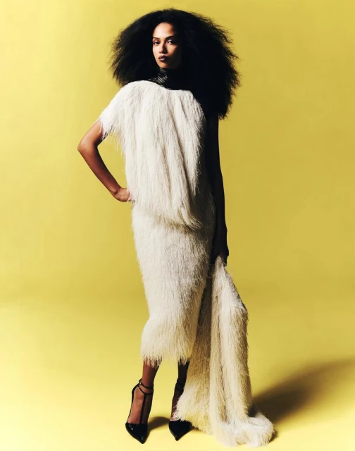 A Black fashion model with an afro hairstyle poses in a stylish, fringed white dress and black high heels against a yellow backdrop.