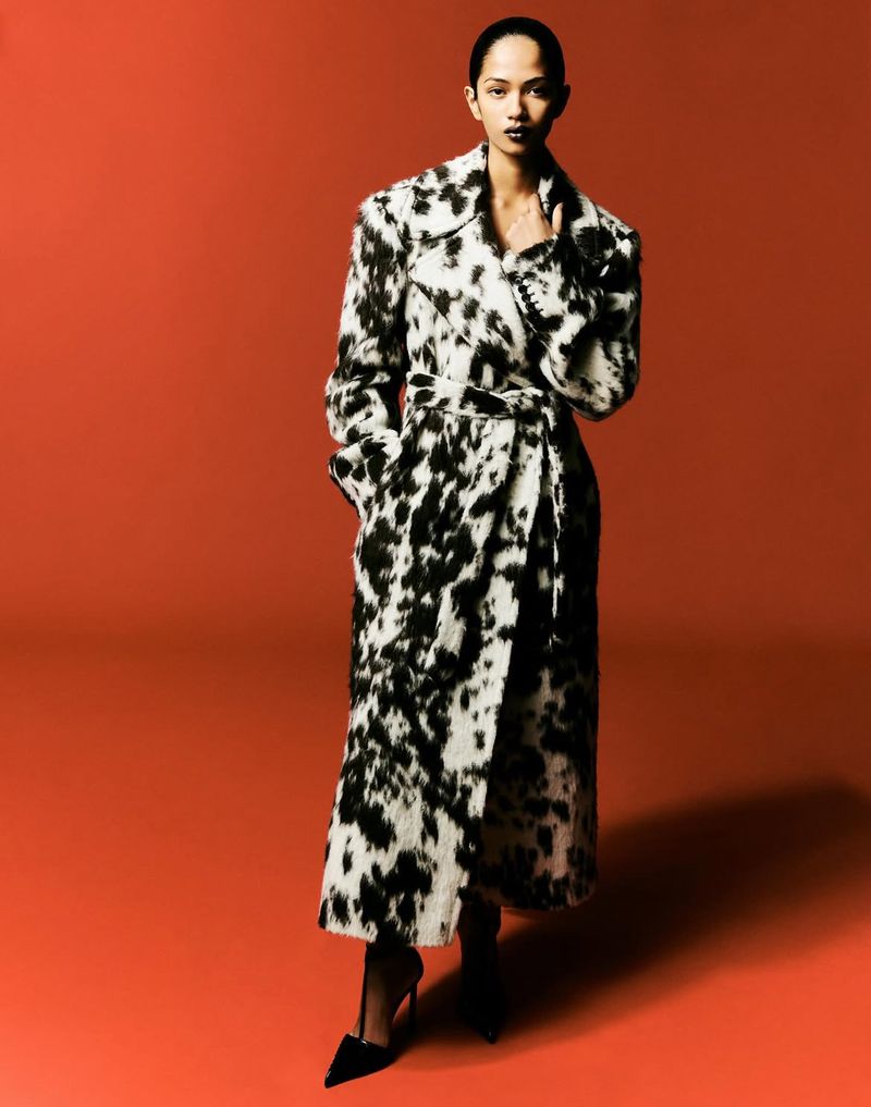 A woman in a black-and-white patterned coat stands against an orange background, showcasing a full-length fashion portrait in studio lighting.