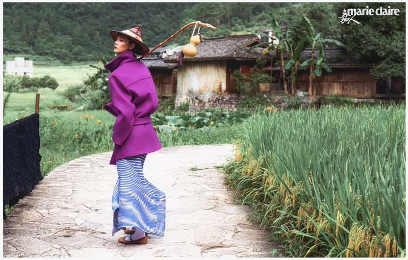 Fashion shot featuring a woman in a purple coat, blue striped skirt, and straw hat, posing on a stone path with rice fields.