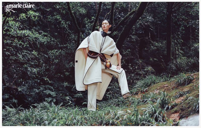 Fashion editorial shot of an Asian woman modeling a cape coat and pants in a lush green forest for Marie Claire magazine.