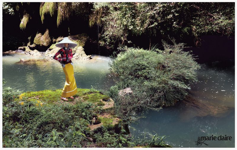 A fashion shot in a lush outdoor setting showcases an adult female in a wide-brimmed hat, patterned jacket, and yellow skirt, posing by a river.