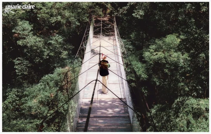 Person in dark jacket and light pants walks across a long suspension bridge surrounded by dense green forest and vegetation.