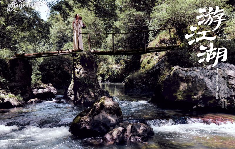 A woman in flowing cream-colored clothing walks along a wooden bridge over a rocky river surrounded by lush green trees and foliage.