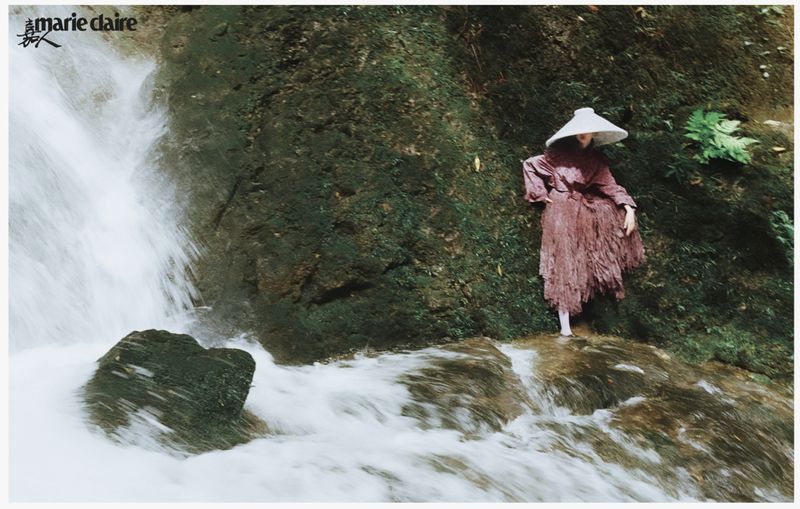 Woman in a long dress and wide-brimmed hat stands on rocks near a waterfall cascading into a pool, surrounded by lush greenery.