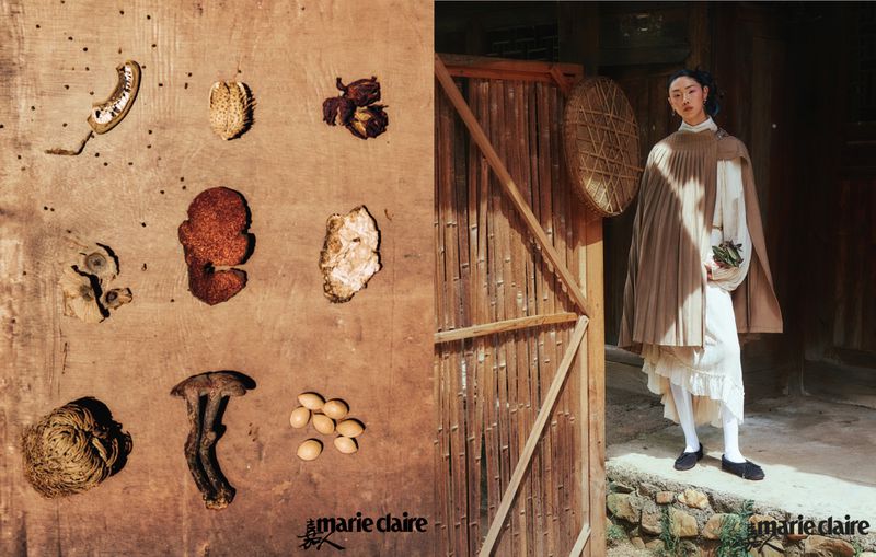 A young Asian woman in a layered white dress and tan cape, stands outdoors next to a bamboo fence; dried botanicals sit on wood paneling.