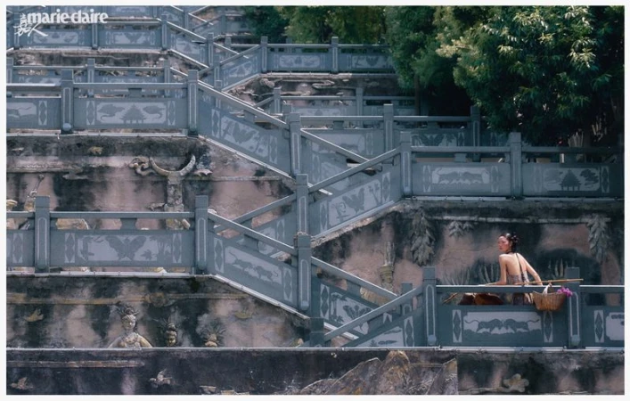 Asian woman in a backless top and straw hat sits on a weathered stone balcony with carved details, overlooking a green, leafy background.