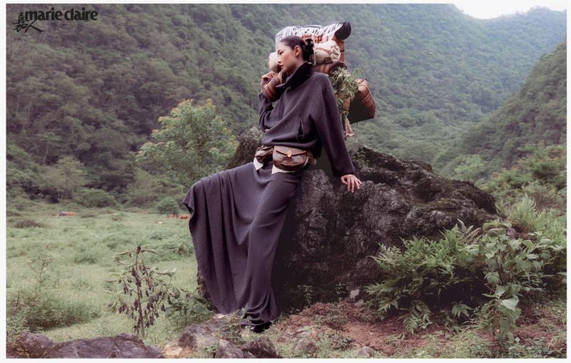 A woman in a long dark outfit sits on a rock in a mountain landscape with a person carrying a load behind her, shot for Marie Claire.