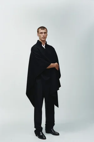 Full-length portrait of a male model wearing a black draped cape and trousers against a pale grey studio background.