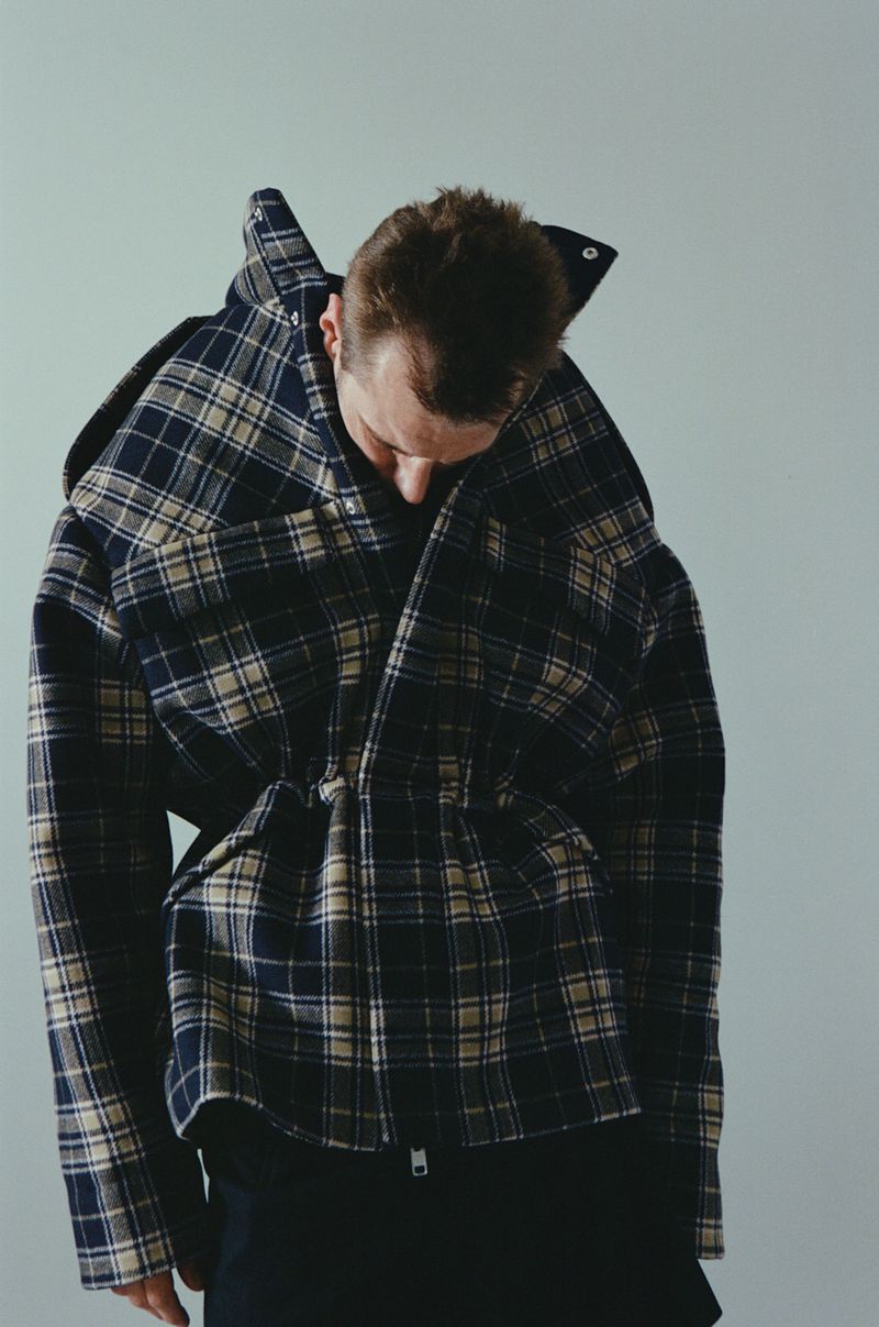 Man in oversized plaid jacket poses indoors against a neutral backdrop, showcasing a modern fashion statement and unique design.