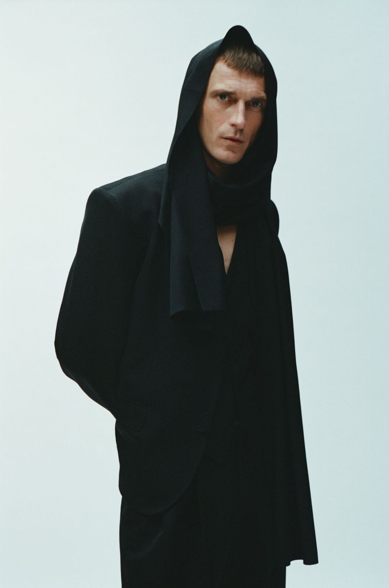 Portrait of a stylish male model wearing a black hooded garment against a neutral backdrop in a fashion editorial style.