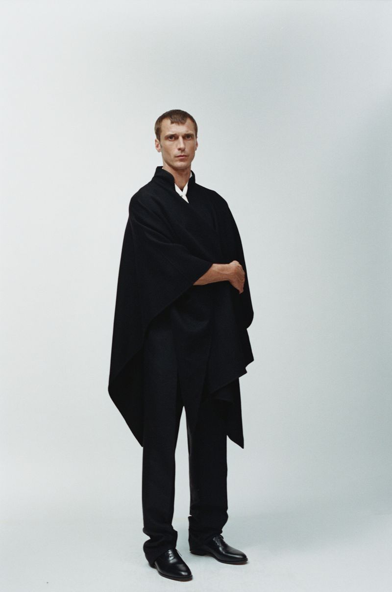Adult male in a full shot wearing a black kimono-style wrap and black dress pants, standing in front of a clean white background.