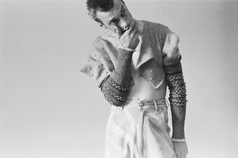 Black and white portrait of a man with a hand to his face, wearing an open sweater and light-colored denim jeans against a plain background.