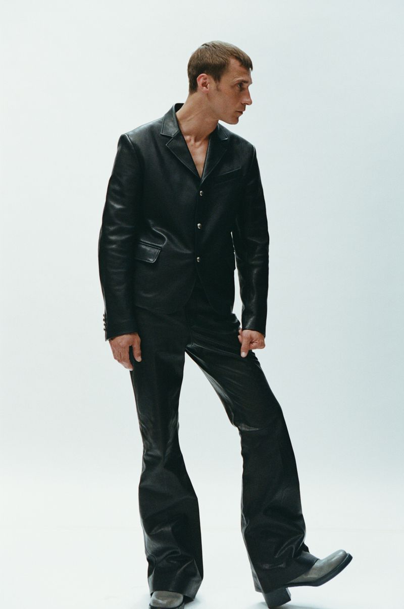 A slender adult male in a black leather suit poses in a brightly lit photo studio, wearing a suit jacket, flared pants, and block-heeled boots.