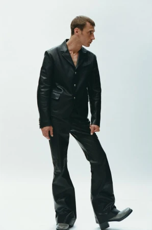 A slender adult male in a black leather suit poses in a brightly lit photo studio, wearing a suit jacket, flared pants, and block-heeled boots.