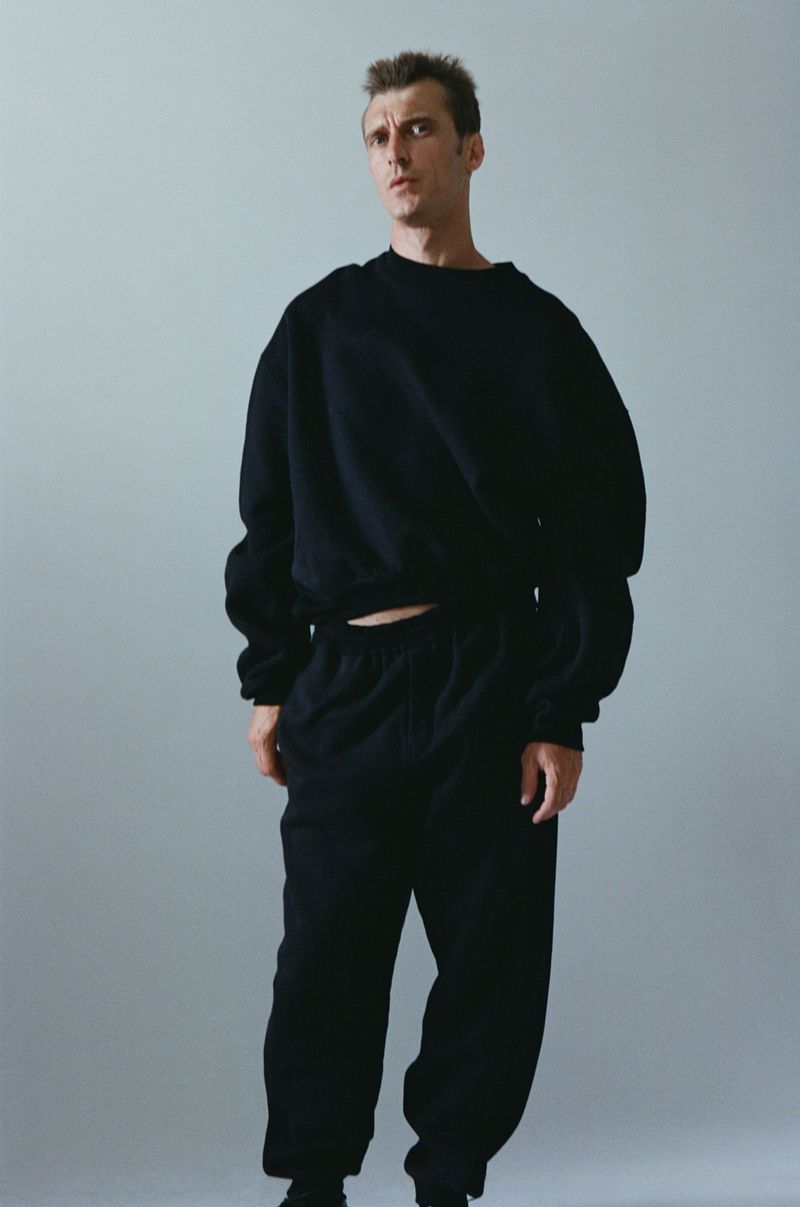 Clement Chabernaud posing in an oversized black sweatshirt and matching sweatpants against a light grey studio backdrop.