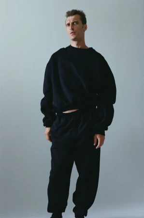 Clement Chabernaud posing in an oversized black sweatshirt and matching sweatpants against a light grey studio backdrop.