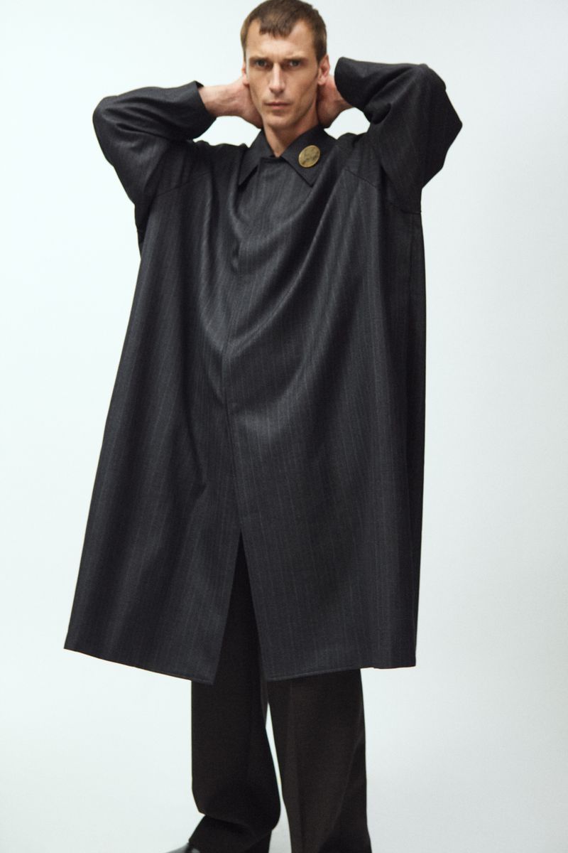 A fair-skinned adult man poses in an oversized black coat with a gold pin, with hands behind his head in a studio setting.
