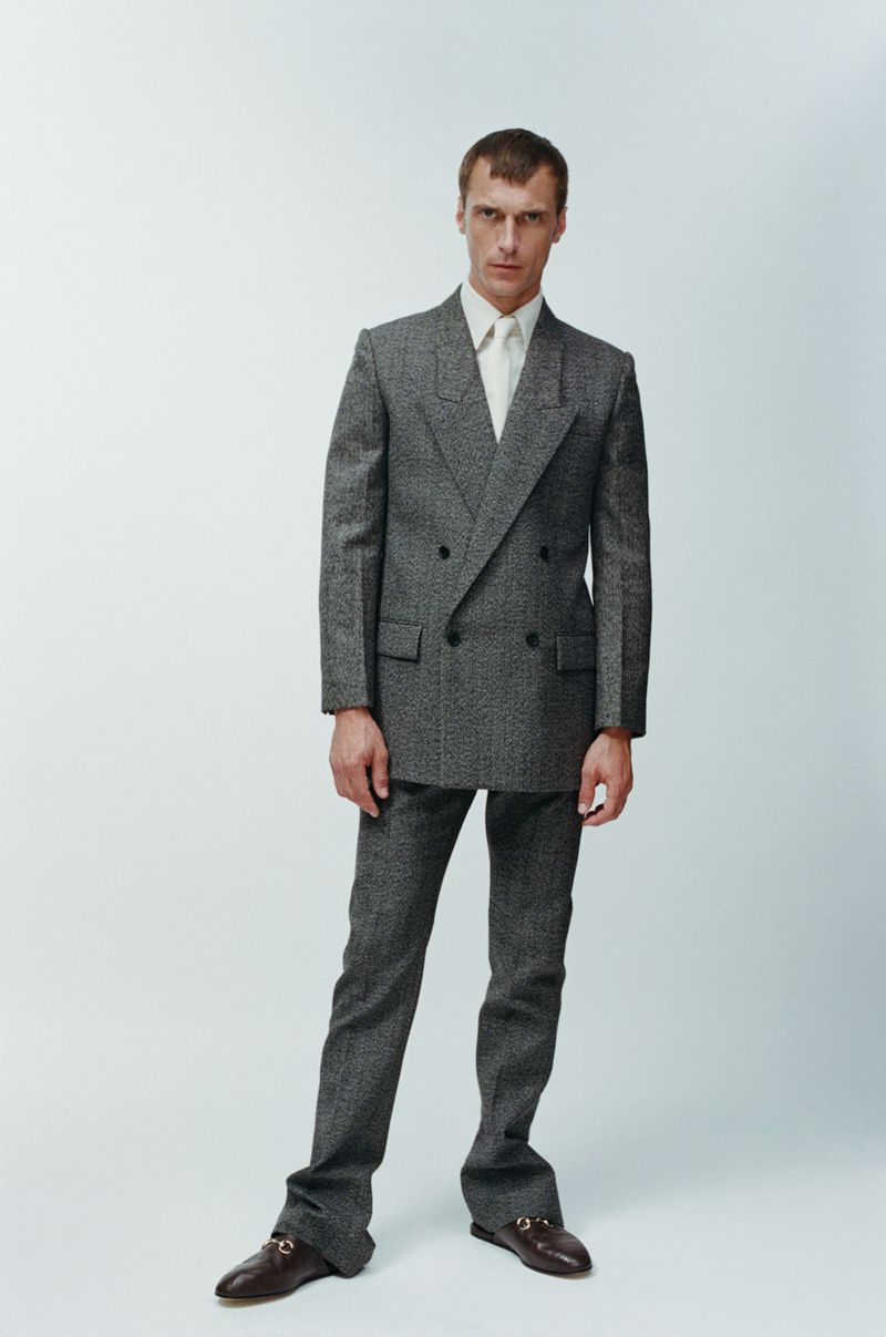 A man in a grey double-breasted suit and dress shoes stands in a neutral studio setting, showcasing contemporary menswear fashion.