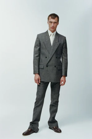 A man in a grey double-breasted suit and dress shoes stands in a neutral studio setting, showcasing contemporary menswear fashion.