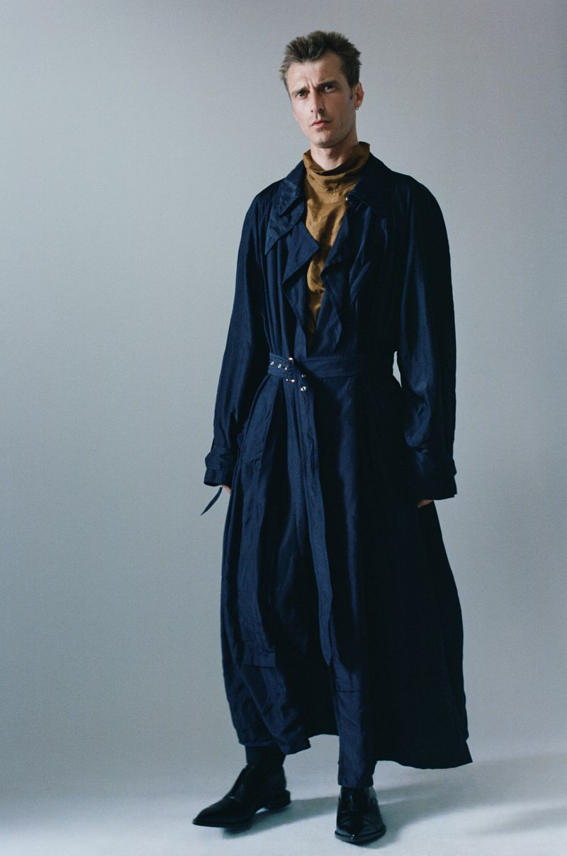 Male fashion model poses in a long blue trench coat and black shoes in a studio setting with a neutral background, looking at the camera.