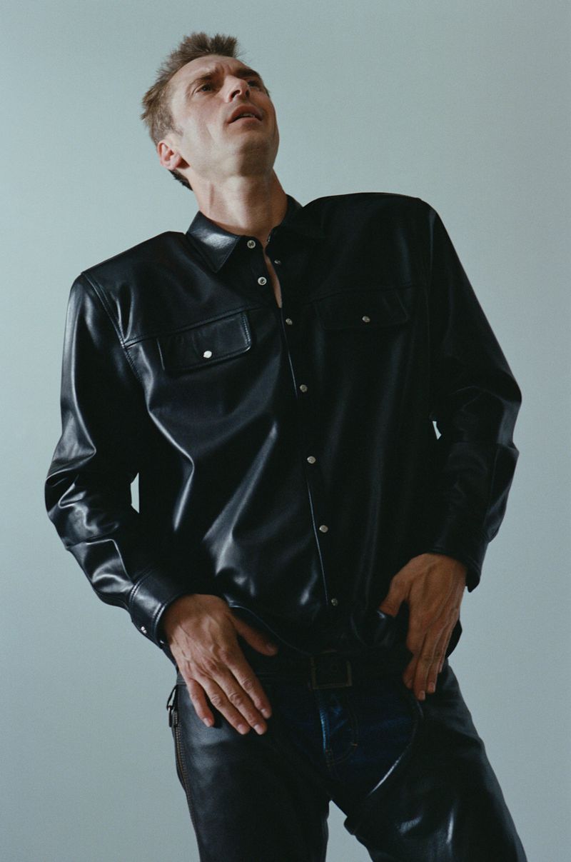 An adult man wearing a black leather shirt and black leather pants poses against a light gray background in a fashion shot.