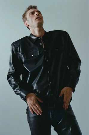 An adult man wearing a black leather shirt and black leather pants poses against a light gray background in a fashion shot.