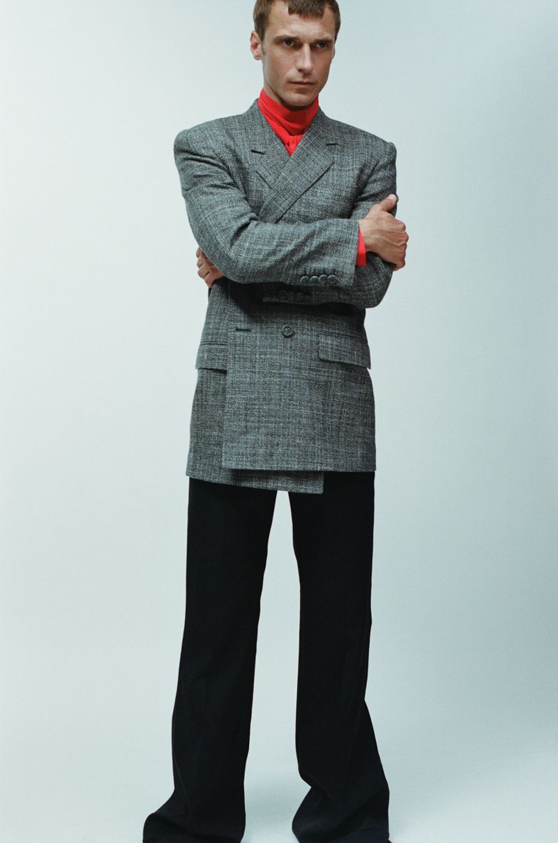 A slender man posing in a gray suit jacket and black pants in a studio setting with soft lighting for a fashion editorial.