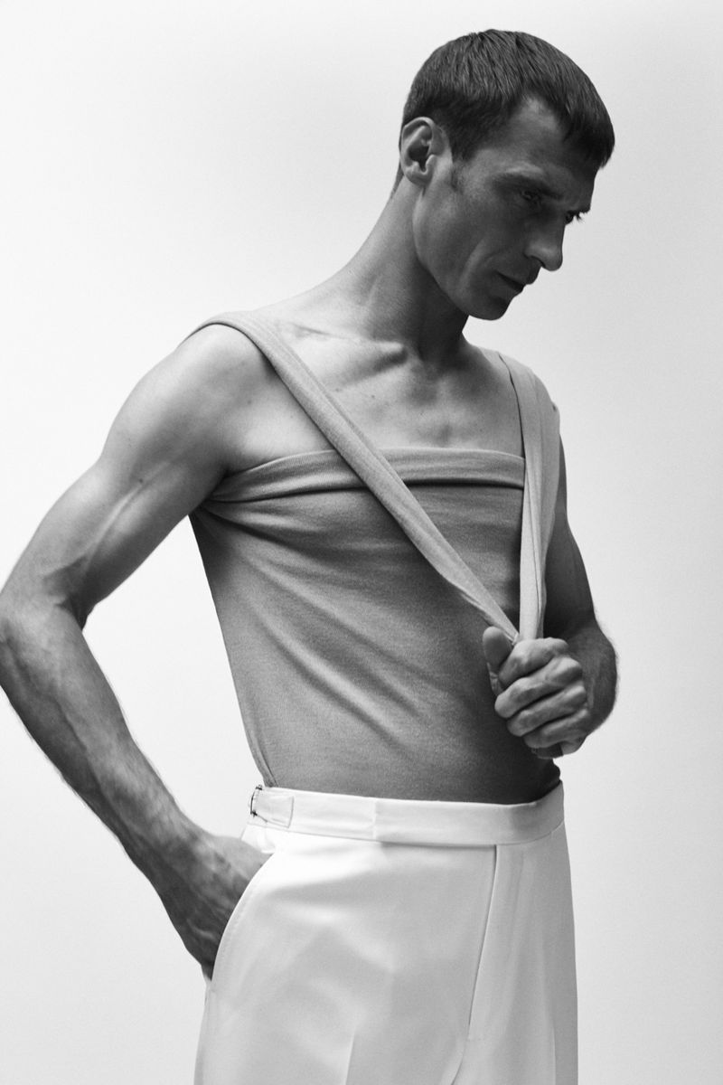Black and white studio portrait of a slim adult man wearing a tank top and white pants, looking down with his hand in his pocket.