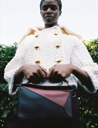 Model wearing a textured white coat and holding a color-blocked handbag in a low-angle shot against a green foliage background.