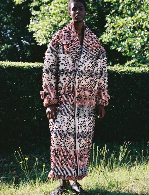Model Onyinyechi Chijioke standing outdoors in front of a lush hedge wearing a long, multi-textured fringed statement coat.
