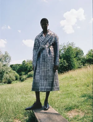 Model standing on a wooden plank wearing a patterned long belted coat against an outdoor grassy meadow and blue sky.