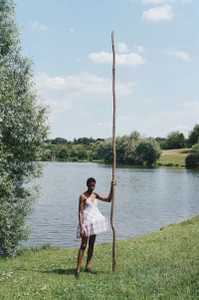Model Onyinyechi Chijioke holding an extremely tall wooden stick while standing in a white patterned mini dress by a lake.