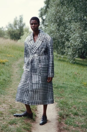 Model Onyinyechi Chijioke standing on a dirt path wearing a long houndstooth patterned coat and black shoes.