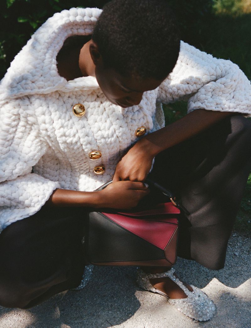 Woman in a white knit jacket with gold buttons, black pants and embellished shoes, holding a burgundy and black geometric handbag outdoors.
